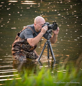 Ontario nature photographer Trevor Pottleburg standing in the water in hip-waders, looking through a camera viewfinder.