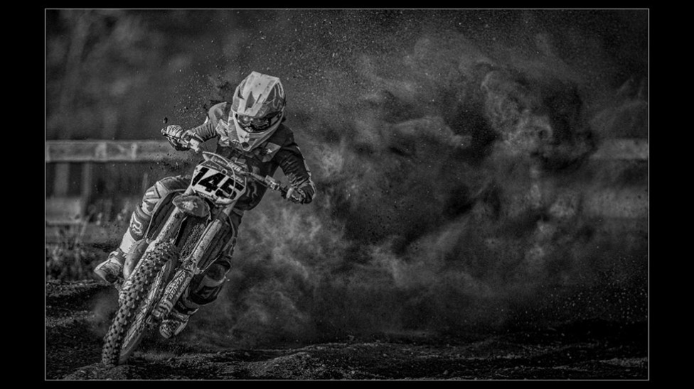 A black and white image of a motocross bike rider with a cloud of dust behind him.