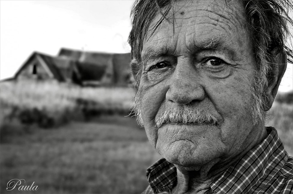 A gritty black and white portrait of a man with a weathered face and a dilapidated building in the background, by photographer Paula Lirette.