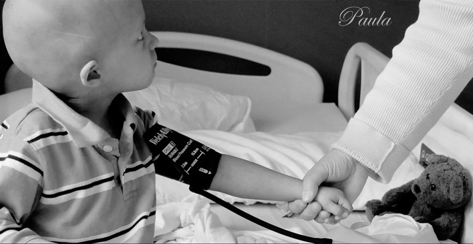 A photo of a bald child with a blood pressure cuff on his arm, holding hands with a person in a white coat. By photographer Paula Lirette.