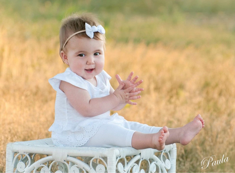 A warm summer scene of a toddler with a bow in her hair, dressed in white. By photographer Paula Lirette.