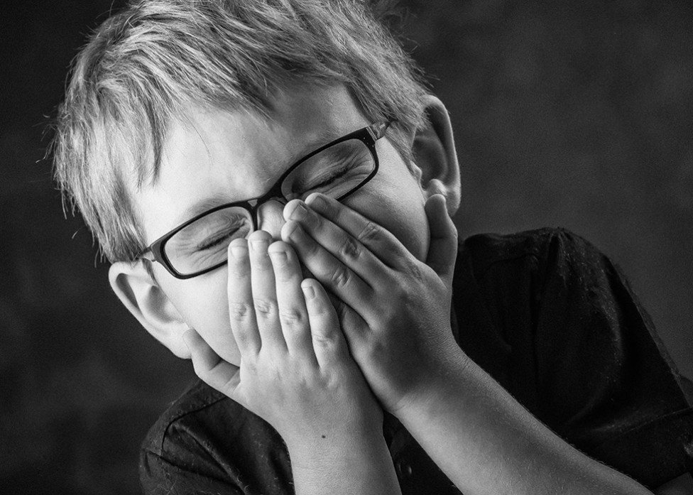 A little boy with glasses giggles shyly behind his hands in this portrait by Louise Vessey